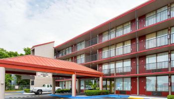 The image shows a multi-story hotel building with a red roof and exterior walkways, and a covered entrance area where a vehicle is parked.