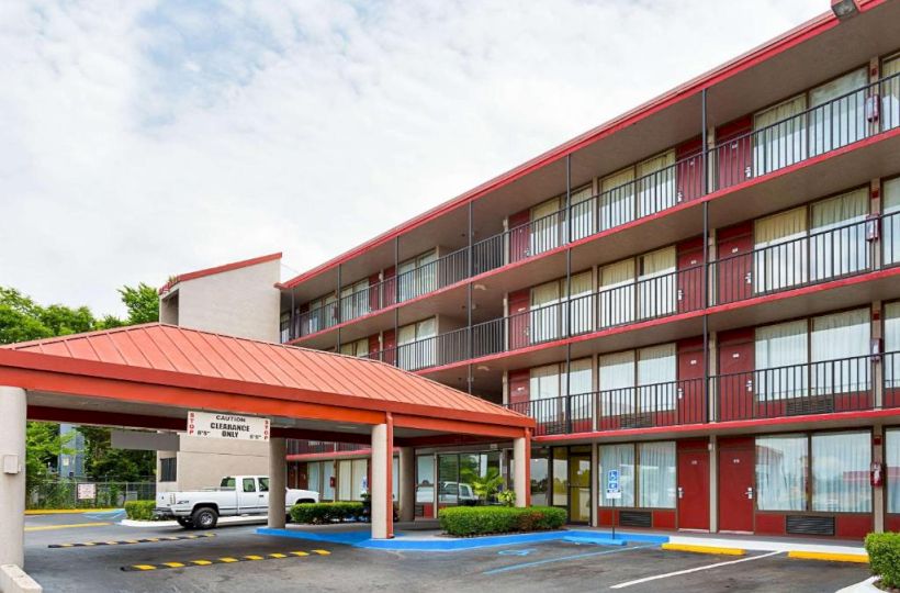 The image shows a multi-story hotel building with a red roof and exterior walkways, and a covered entrance area where a vehicle is parked.