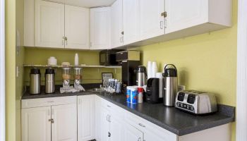 A small kitchen or break room with white cabinets, a coffee station, toasters, and various cups and containers on a black countertop, under a pale green wall.