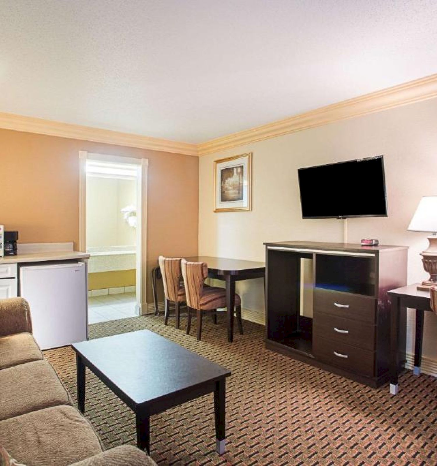 A hotel room with a sofa, coffee table, TV, desk, chairs, lamps, mini-fridge, and microwave. Beige walls and carpet, simple and cozy decor.