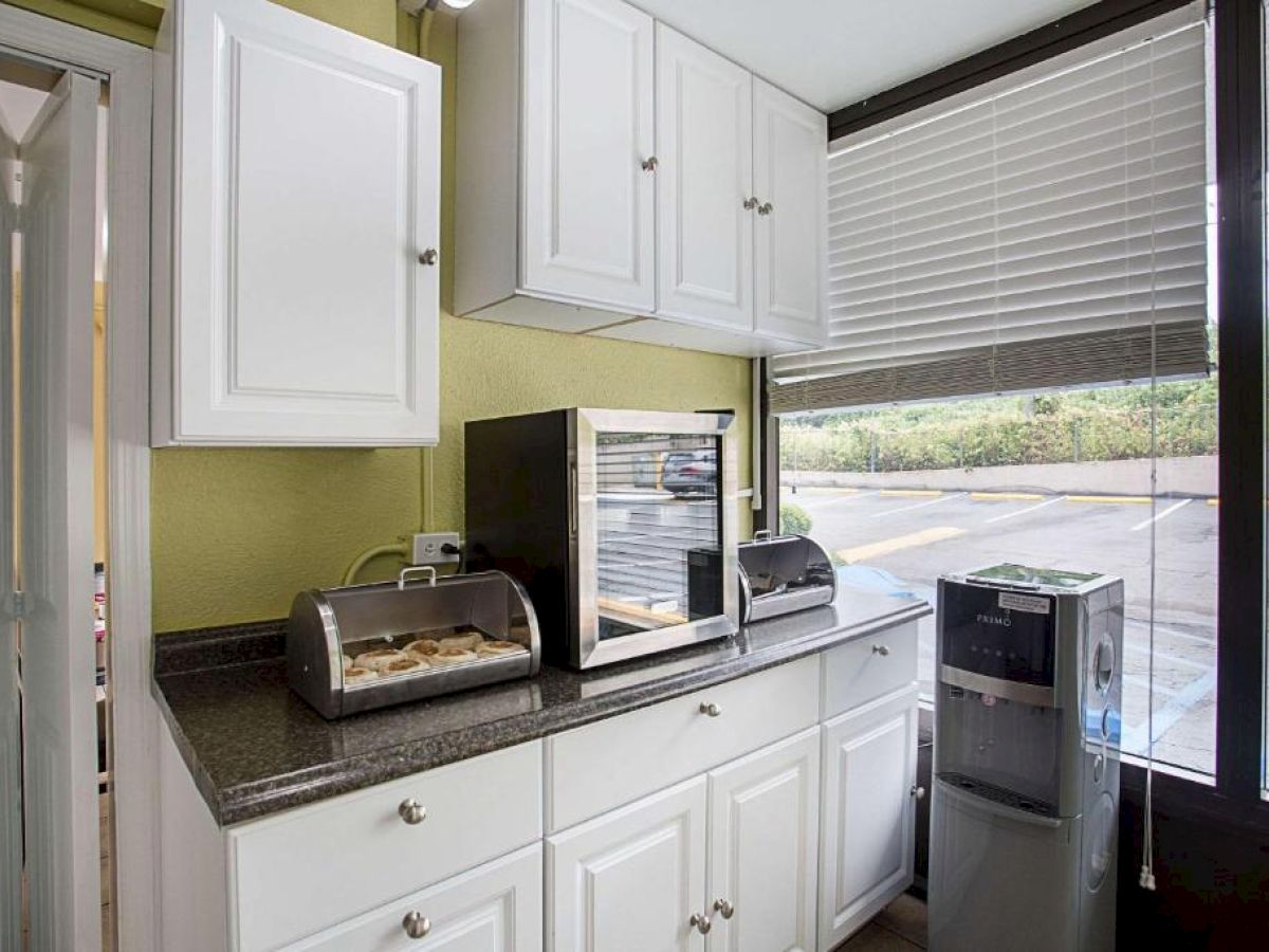 A kitchen area with white cabinets, countertop appliances, a food display, and a water cooler by a large window with blinds, is in the image.