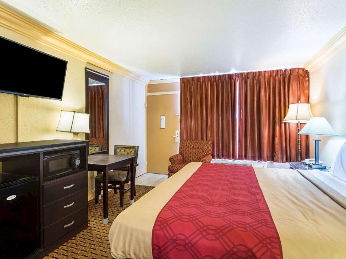 A hotel room features a bed with red and beige bedding, a TV above a dresser, table with chairs, lamps, a window with curtains, and an armchair.