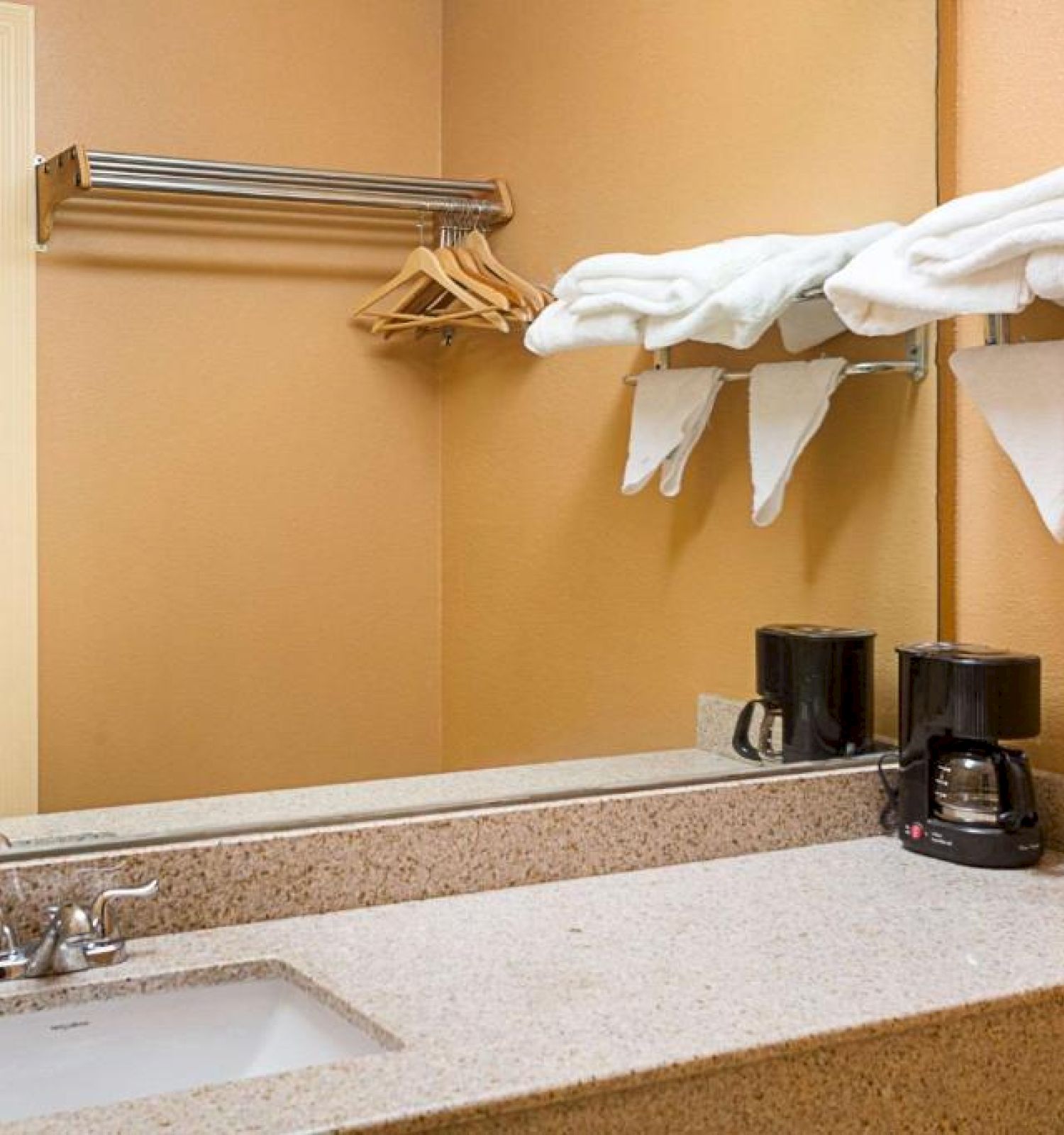 A bathroom sink with a large mirror, towels on a rack, a coffee maker, and clothes hangers. There is a doorway leading to another room.