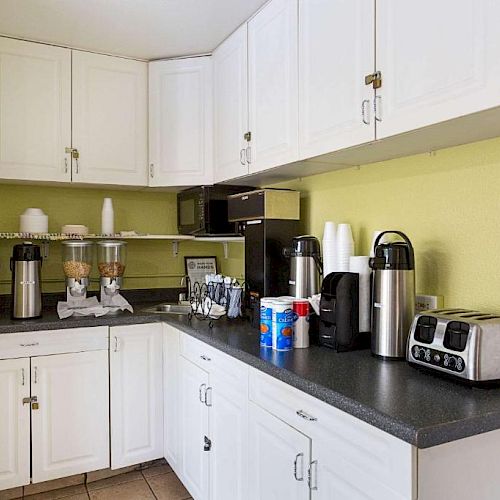 A small kitchen area with white cabinets, coffee dispensers, a toaster, disposable cups, a microwave, and a can of coffee on the countertop.