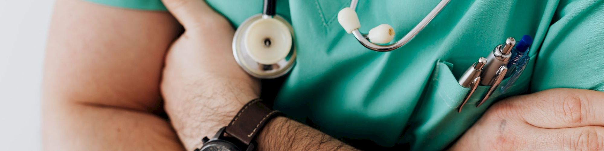 A person wearing green scrubs with a stethoscope around their neck and arms crossed, a watch, and pens in the chest pocket.