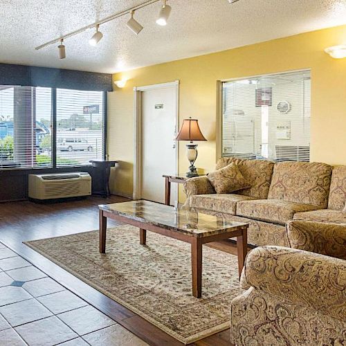 This image shows a cozy living room with patterned sofas, a glass coffee table, a lamp, a framed picture on a yellow wall, and large windows.