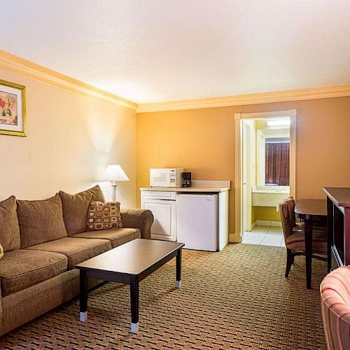 A cozy hotel room with a sofa, coffee table, desk, chairs, flat-screen TV, mini-fridge, microwave, and wall art; a bathroom in the background.