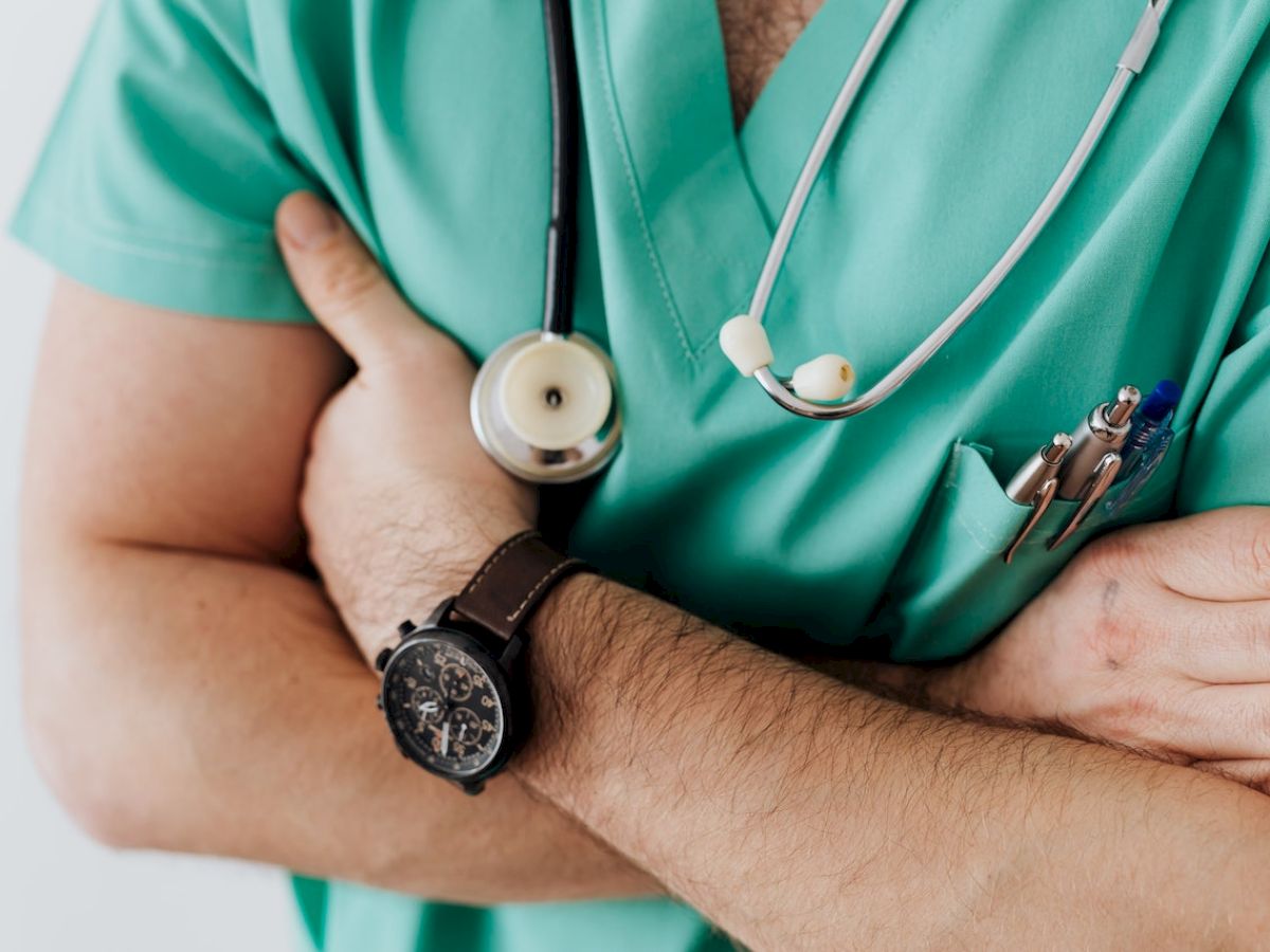 The image shows a person in green scrubs with a stethoscope around their neck, arms crossed, and a watch on their wrist, holding pens in their pocket.