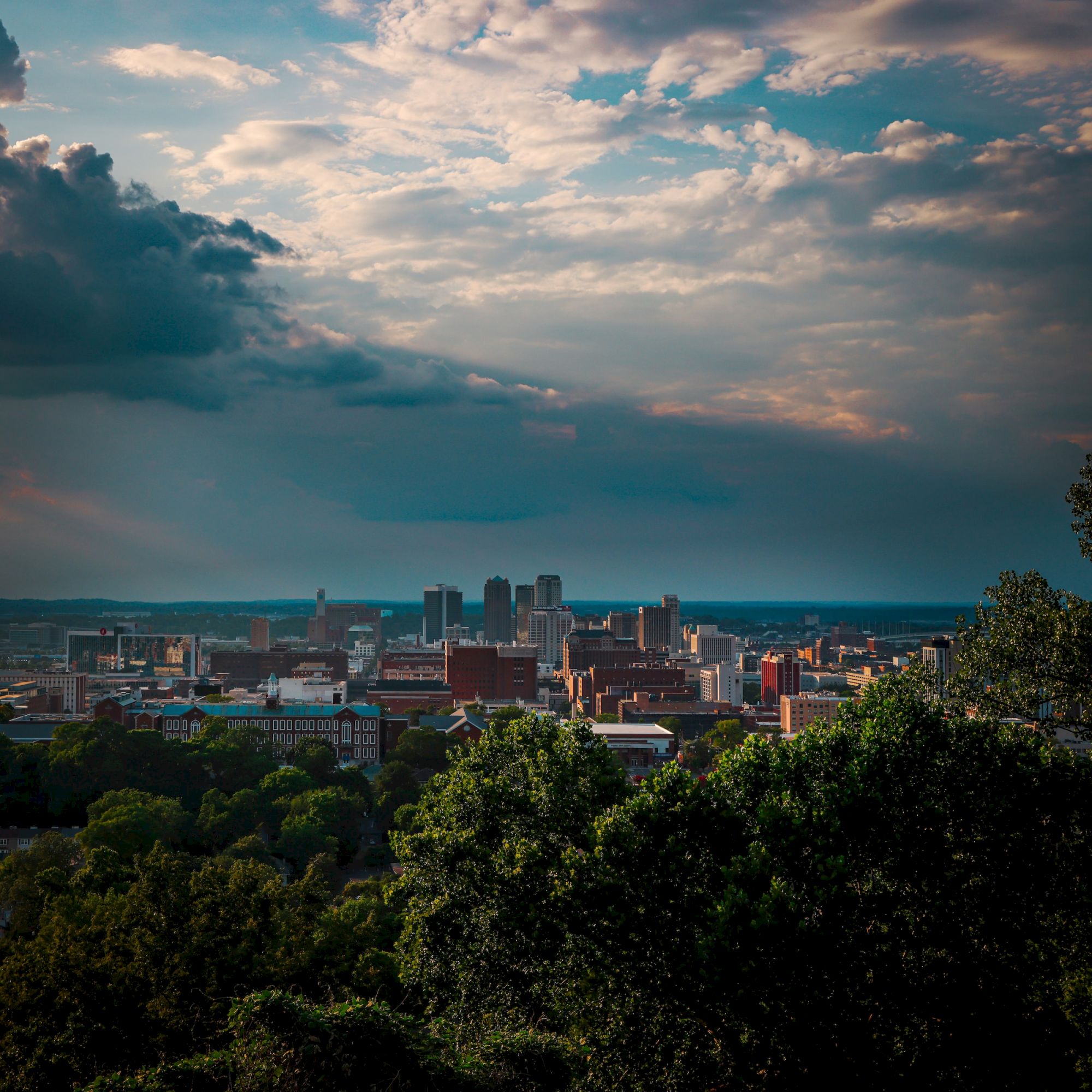 The image shows a city skyline with several buildings under a dramatic sky, viewed from a distance above treetops, giving a scenic and picturesque perspective.