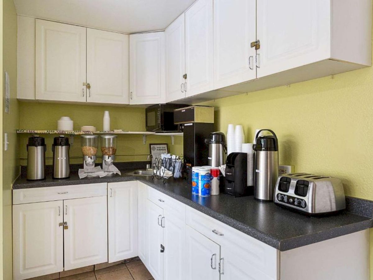 A small kitchen space with white cabinets, coffee dispensers, a toaster, disposable cups, various silverware on the countertop, and microwaves.