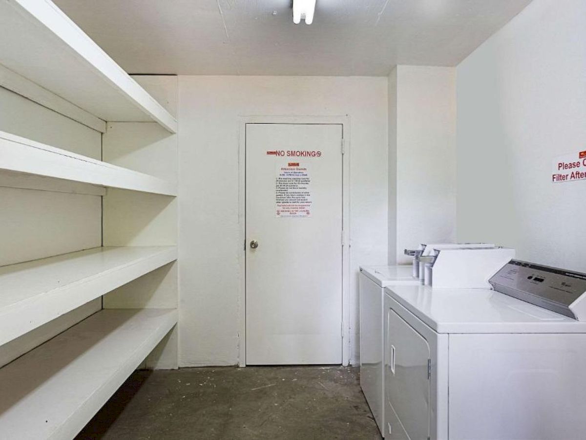 The image shows a laundry room with empty shelves on the left, a door in the center, and a washing machine on the right with a sign on the wall.