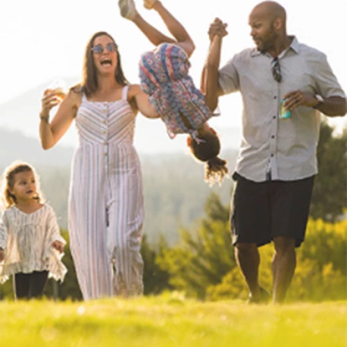 A family is walking outdoors in a sunny setting. The parents hold hands with two children, one of whom is being lifted into the air, smiling.
