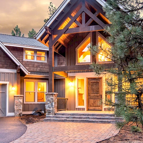 The image depicts a beautifully lit, large modern house with a three-car garage and wooden exterior, surrounded by trees during twilight.