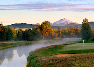 A serene landscape with a golf course, a river, trees, and a snow-capped mountain in the background, under a clear sky.