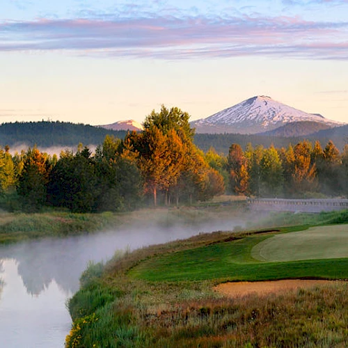 A serene landscape with a golf course, a river, trees, and a snow-capped mountain in the background, under a clear sky.