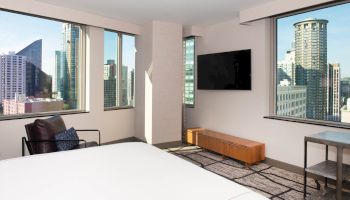 A modern hotel room with large windows showcasing a city view, a bed, a TV on the wall, and a chair.