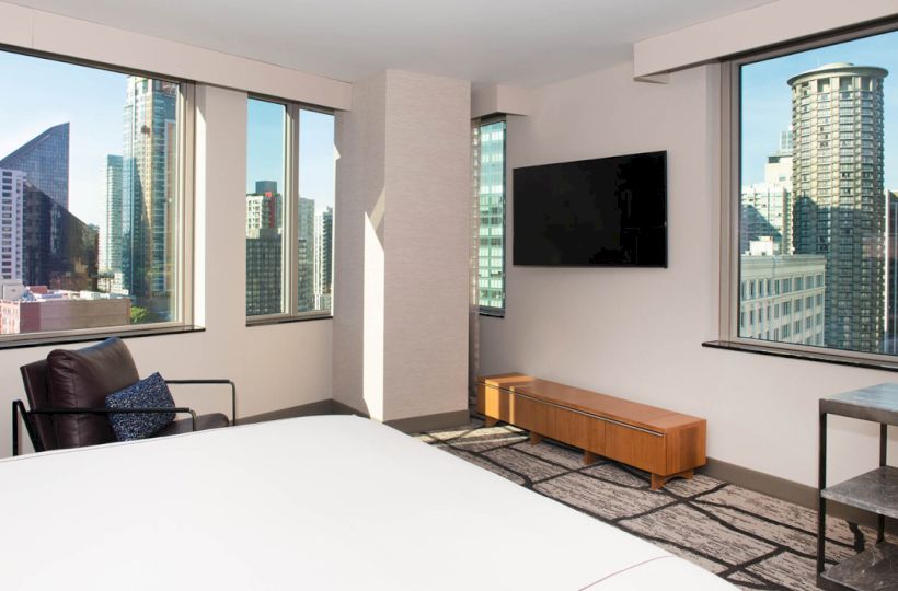 A modern hotel room with large windows showcasing a city view, a bed, a TV on the wall, and a chair.