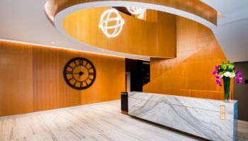 A modern lobby with a sleek, marble reception desk, spiral wooden staircase, large decorative clock on the wall, and floral arrangement.