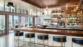 A modern bar with a wooden counter, four blue and gold stools, a well-stocked shelf of liquor bottles, and large windows offering a city view.