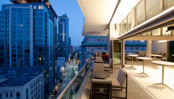 A modern balcony at dusk with chairs and tables, overlooking a lively city street with tall buildings and illuminated windows, in an urban setting.