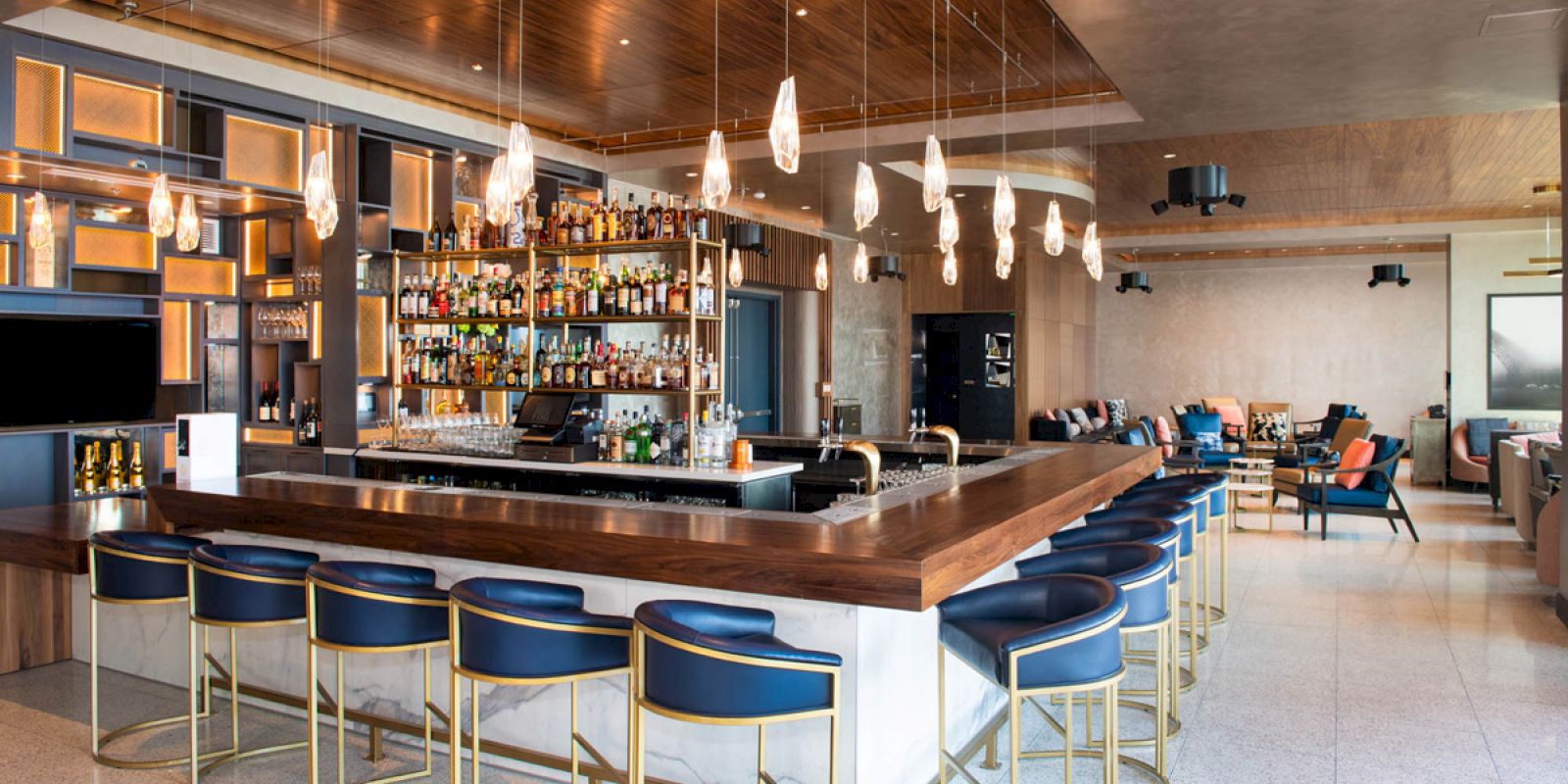 The image shows a modern bar with a wooden counter, blue bar stools, hanging lights, and a well-stocked liquor shelf in a spacious area.
