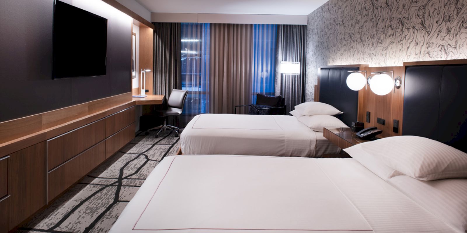 A modern hotel room with two neatly made beds, a wall-mounted TV, a desk and chair, ambient lighting, and a window with curtains.