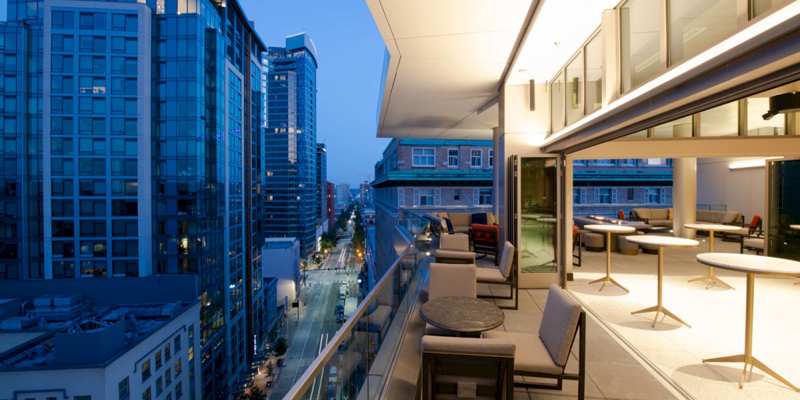 A modern balcony with seating overlooks a city street lined with tall buildings, illuminated as night falls. The interior is well-lit and inviting.