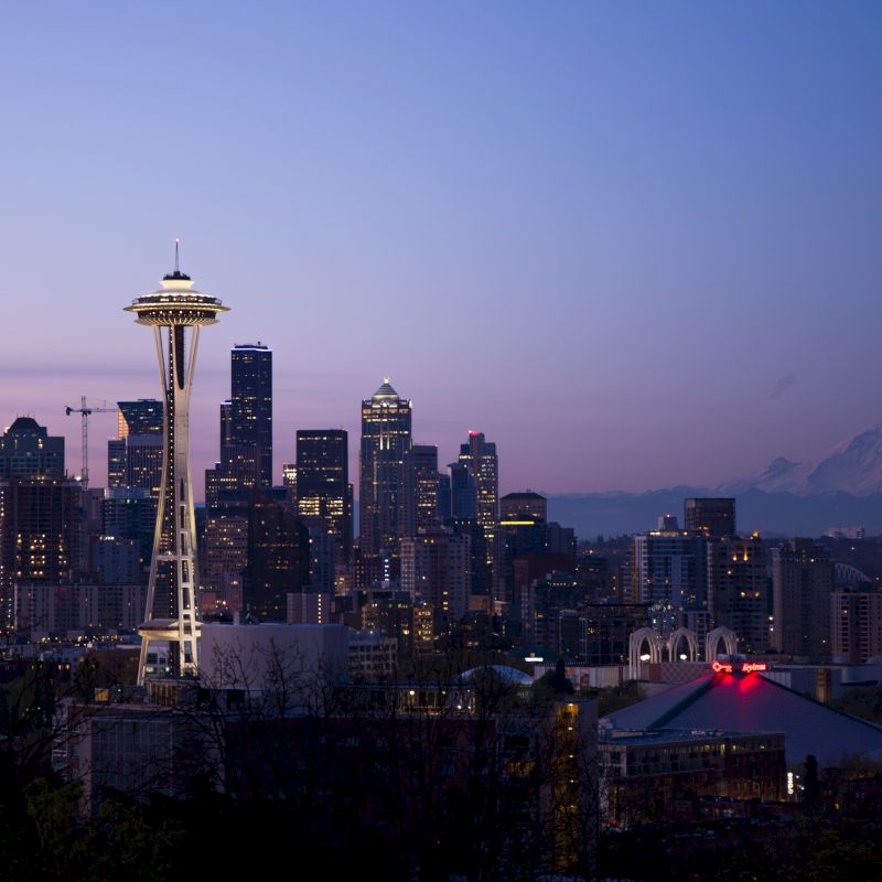 A cityscape at dusk featuring a prominent tower and a skyline of high-rise buildings with a mountain visible in the background.