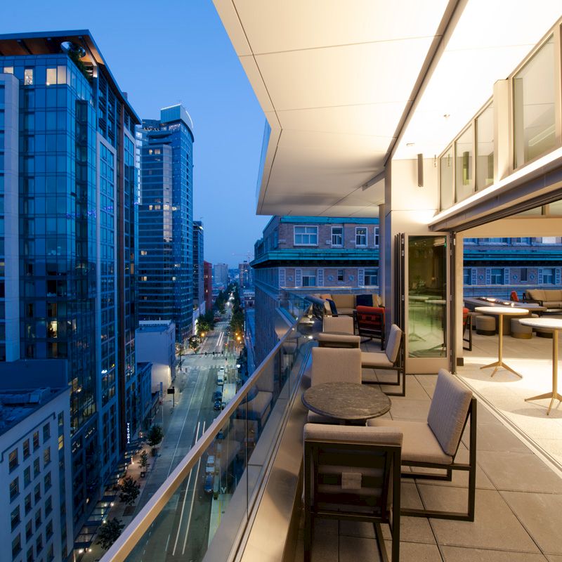 A modern cityscape at dusk with high-rise buildings and illuminated streets, viewed from a sleek balcony featuring seating and tables.