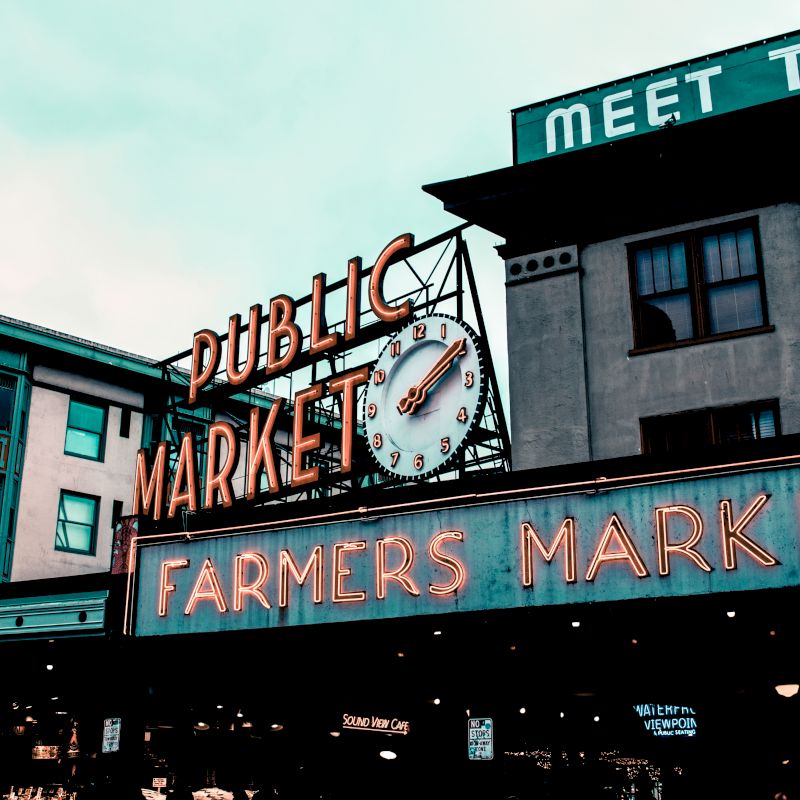 The image shows the iconic entrance sign for a public market and farmers market, with nearby buildings displaying various other signs.