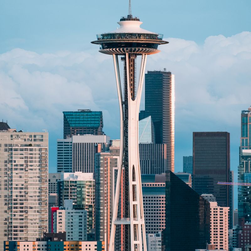 The image shows the Space Needle, a prominent observation tower in Seattle, with city skyscrapers in the background against a partly cloudy sky.