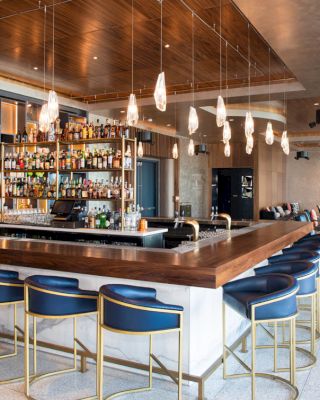 The image shows a modern bar with a large counter, blue bar stools, and shelves stocked with drinks. There is stylish lighting and seating areas.