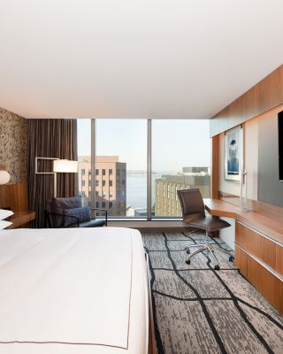 A modern hotel room with a large bed, desk, TV, and a view of city buildings through large windows is shown.