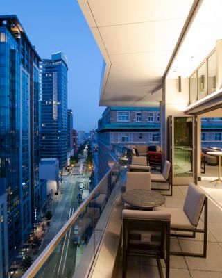 An evening view from a modern balcony overlooking a city street, featuring outdoor seating and tall glass buildings reflecting city lights.