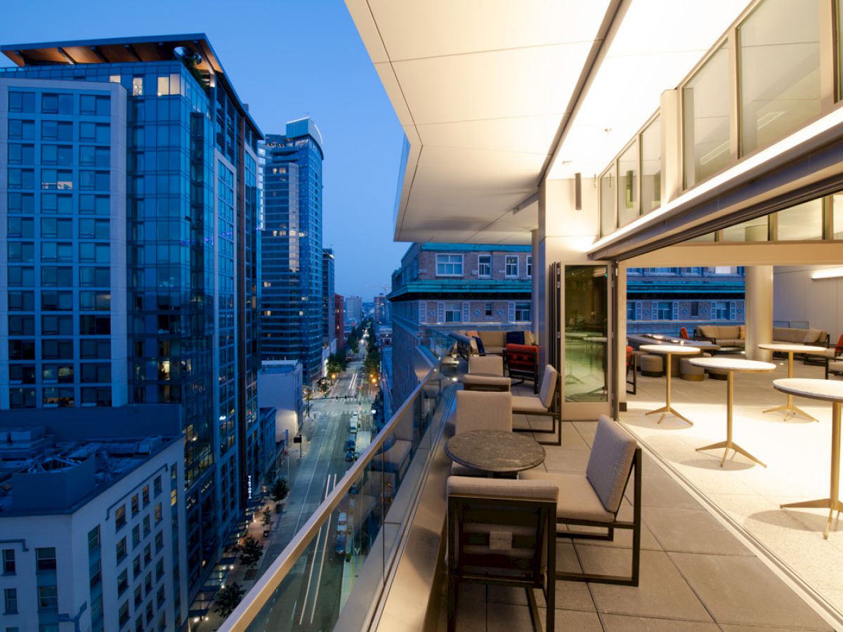 Cityscape view with modern buildings at dusk, seen from a balcony with seating and tables on the right side.