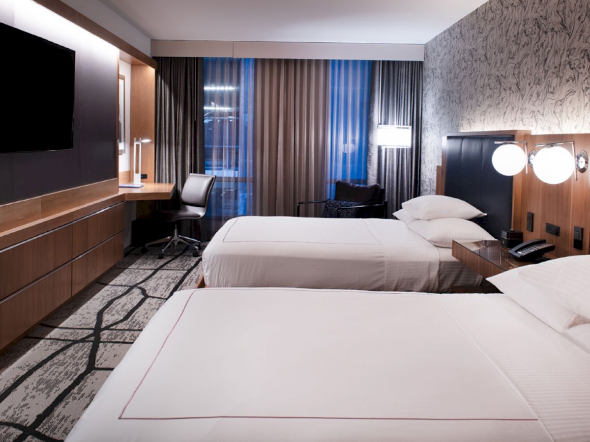 A modern hotel room with two neatly made beds, a wall-mounted TV, a desk with a chair, and contemporary decor.
