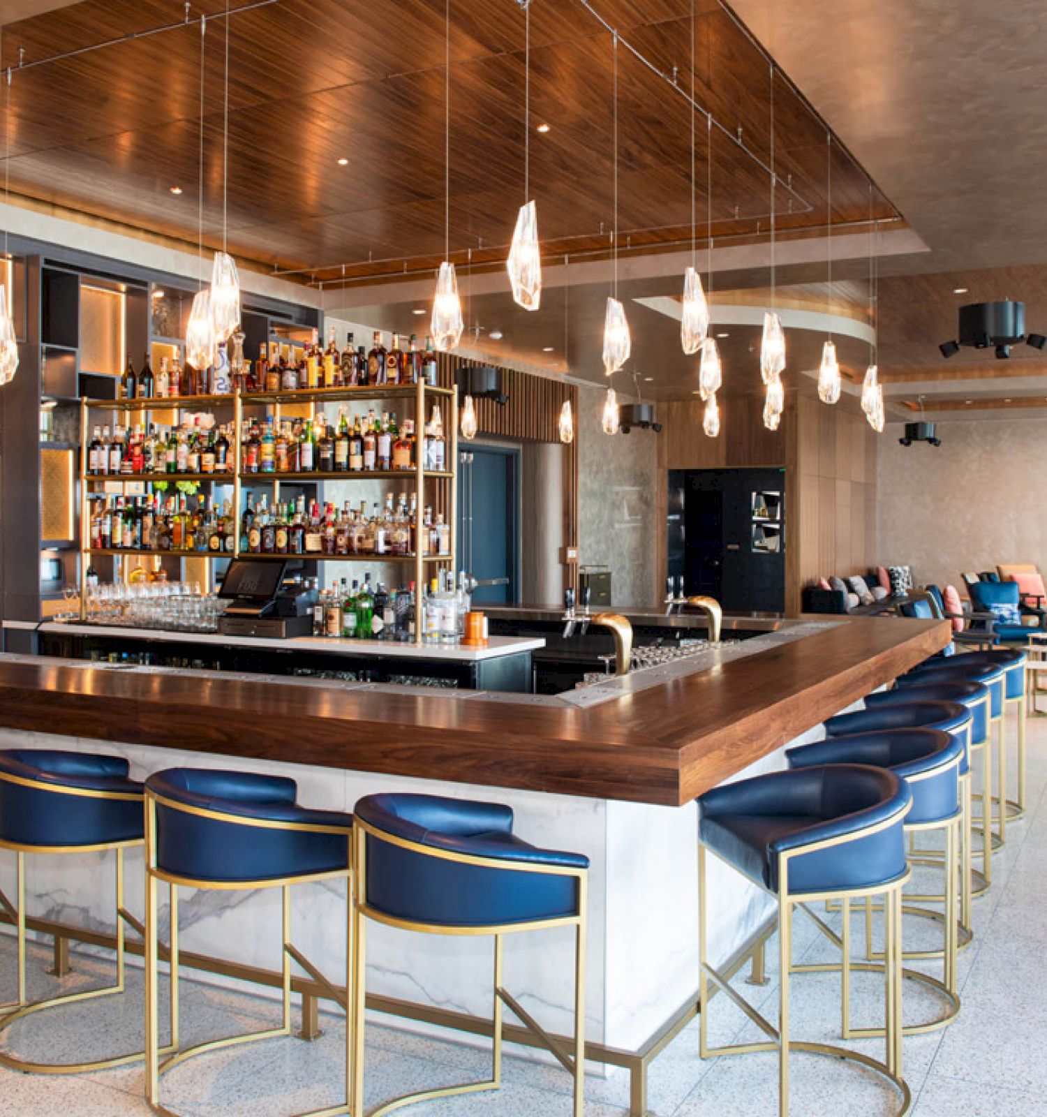 A modern bar with blue and gold stools, a stocked back bar, hanging lights, and a lounge area with chairs and a TV in the background.