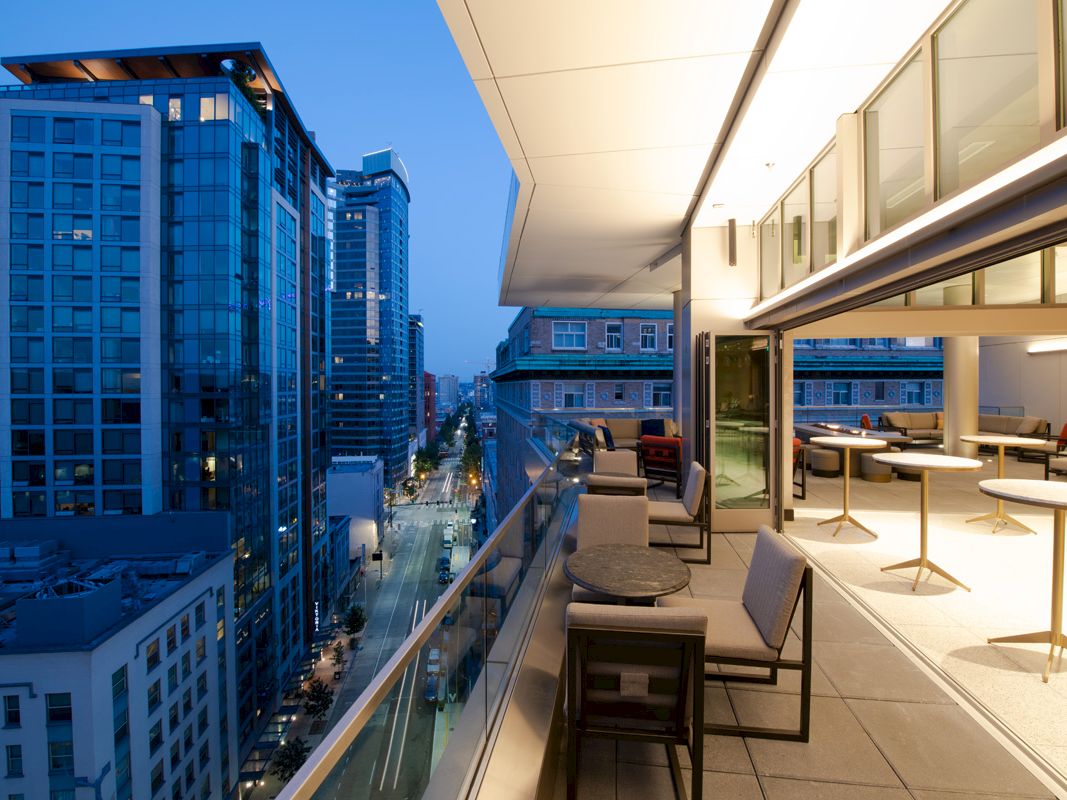 A modern cityscape at dusk featuring high-rise buildings and a balcony with seating and tables illuminated by warm indoor lighting.