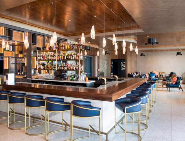 A modern bar with blue cushioned barstools, pendant lighting, and a fully stocked bar, in a spacious lounge area with seating and a TV.