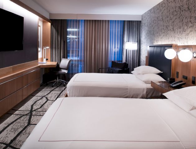 A modern hotel room with two neatly made beds, a flat-screen TV, a desk with a chair, and large windows with curtains.