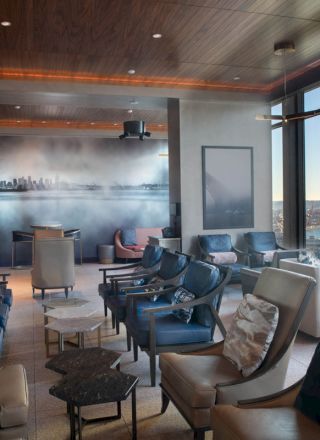 The image shows a modern lounge with comfortable chairs, large windows offering a scenic view of a waterfront with a Ferris wheel, and warm lighting.