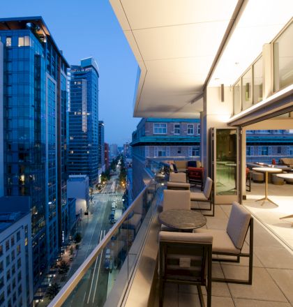 The image shows a modern urban balcony overlooking a city street with tall buildings, featuring outdoor seating and tables under warm lighting.