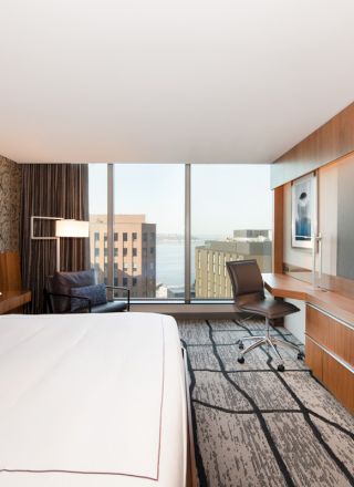 A modern hotel room with a large bed, desk, chair, and big screen TV, offering a cityscape view through a wide window.