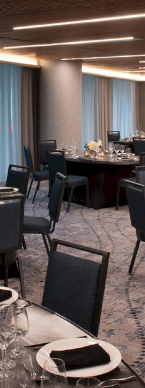 A modern banquet hall is set up with round tables, black chairs, and elegant table settings including flowers and candles, illuminated by overhead lighting.
