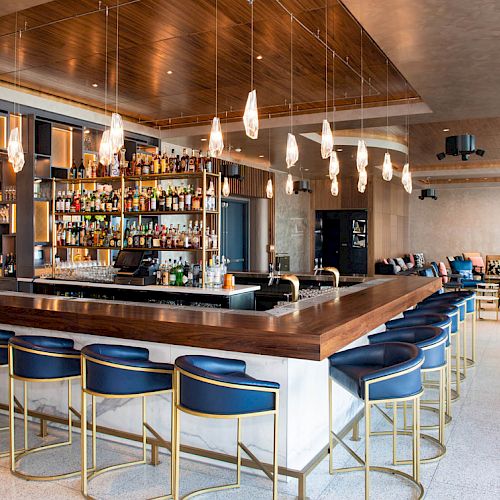 The image shows a modern, stylish bar with blue barstools, a well-stocked bar, hanging lights, and a comfortable seating area.