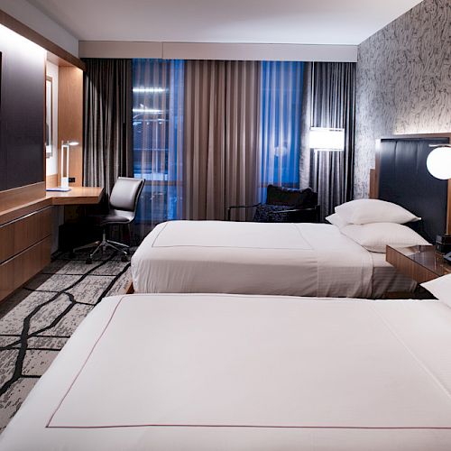 A modern hotel room with two neatly made beds, a flat-screen TV, a desk with a chair, and stylish decor, featuring a large window with curtains.