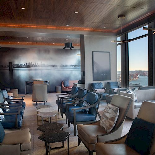 Spacious modern lounge with plush seating, floor-to-ceiling windows showcasing a coastal view, and a cityscape mural on the back wall.