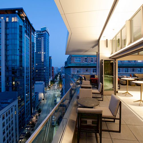 A modern rooftop patio with seating areas overlooks a city street lined with tall buildings as dusk settles in.