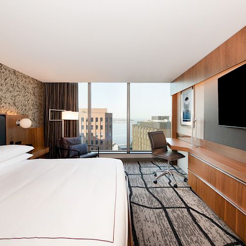 The image shows a modern hotel room with a large bed, wall-mounted TV, desk, chair, and floor-to-ceiling windows offering a cityscape view.