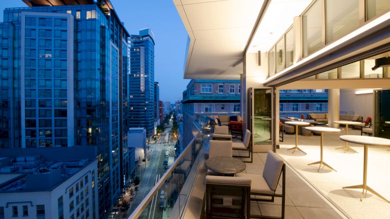 This image shows a modern cityscape with tall buildings, a lit-up street below, and a high-rise balcony with outdoor furniture and tables.